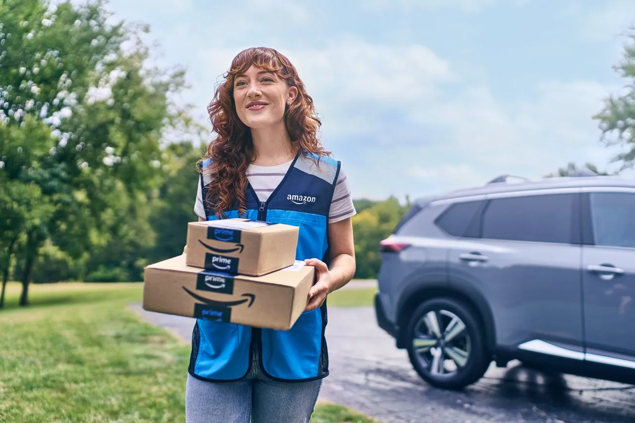 A smiling young woman holds two packages and walks beside a lawn. She wears a blue Amazon vest.