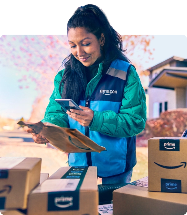 A woman in a blue Amazon vest scans a package with her phone. Around her are half a dozen more packages.