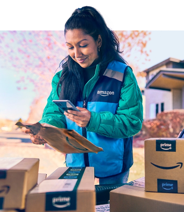 A woman in a blue Amazon vest scans a package with her phone. Around her are half a dozen more packages.