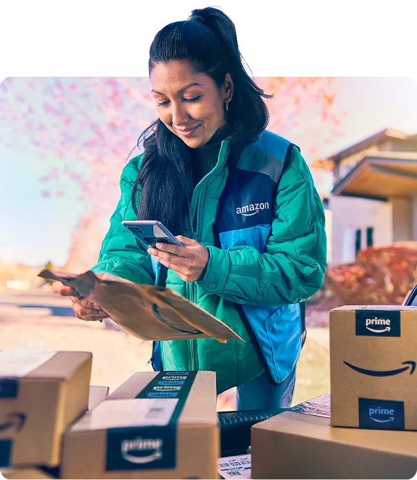 A woman in a blue Amazon vest scans a package with her phone. Around her are half a dozen more packages.