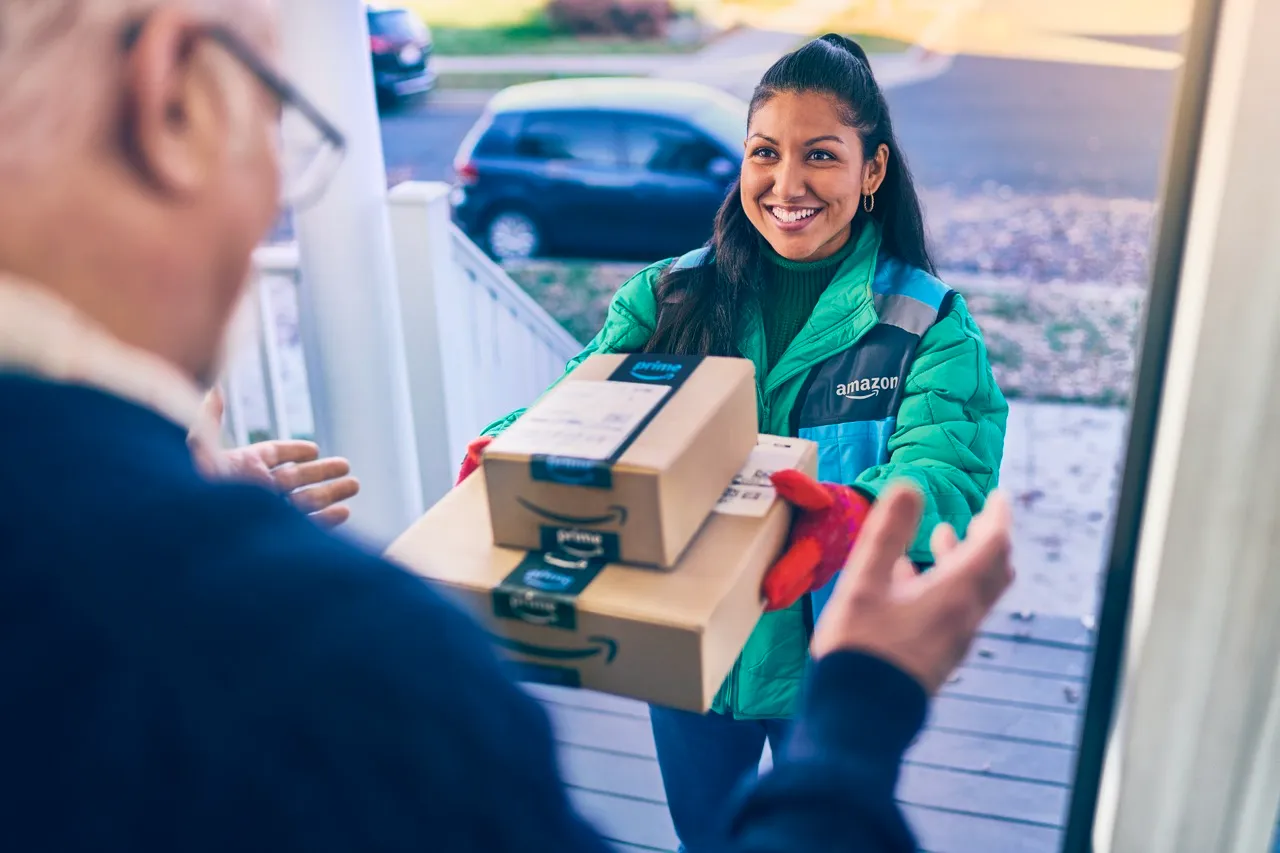 A woman in a blue Amazon vest smiles and hands a man two packages.