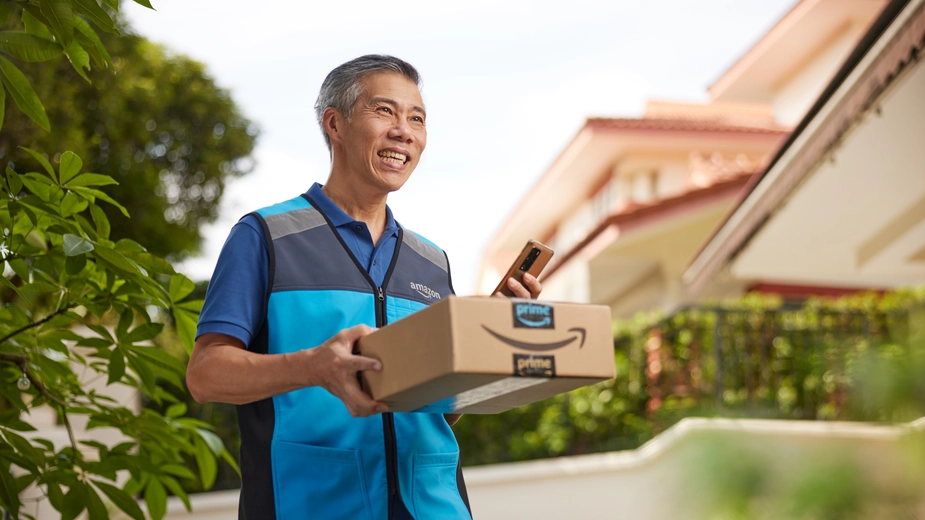Amazon delivery partner in blue uniform holding package and phone smiling outdoors in residential neighborhood