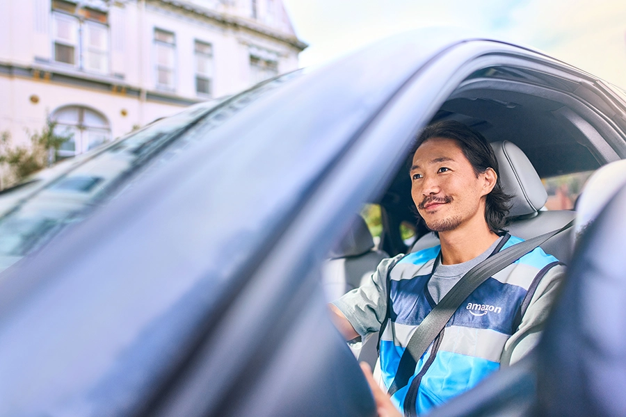 Amazon delivery driver wearing blue safety vest smiling while seated in vehicle with seatbelt fastened.