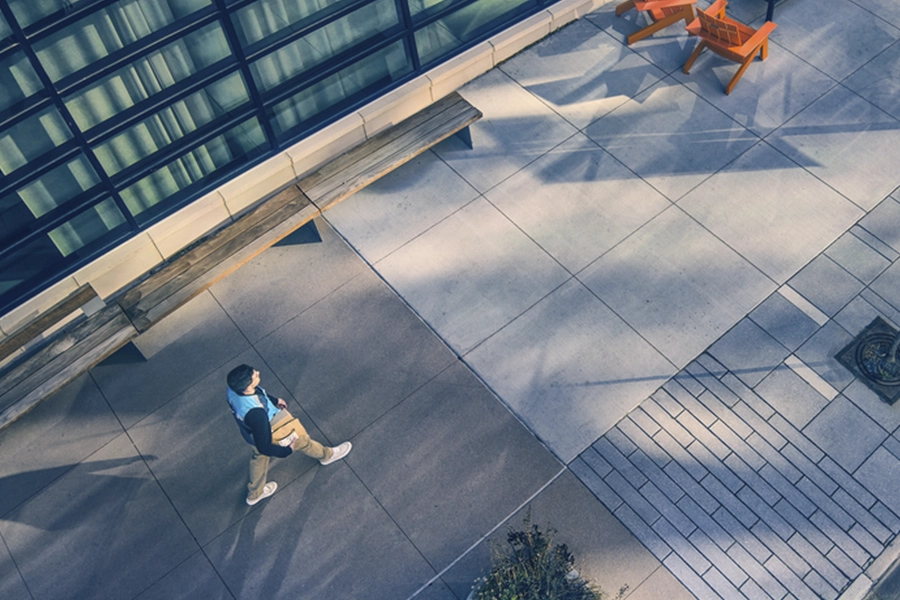 Aerial view of person holding a package walking on an urban street with orange chairs, large tiles, and glass building facade