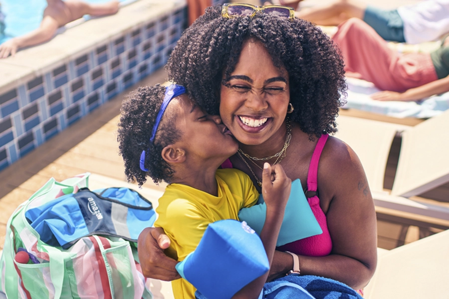 Child kissing mother's cheek poolside, both smiling and holding colorful beach bags and towels on sunny day