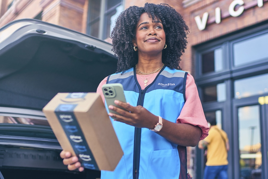 Amazon delivery driver in blue vest holding phone and package by open car trunk outside urban storefront