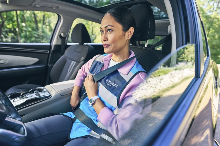 Woman wearing Amazon-branded vest in car driver's seat, putting on her seatbelt while parked on tree-lined street