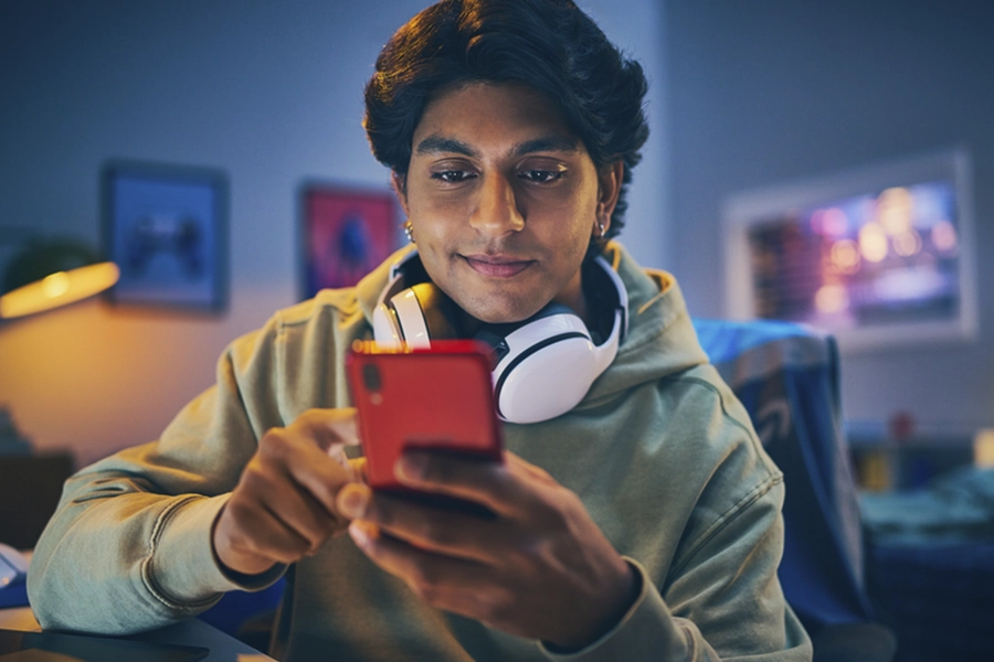  Man in a hoodie with headphones around his neck sitting in a bedroom looking at his phone
