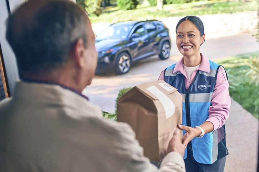 Woman in a blue vest handing an Amazon package to a man at his doorstep